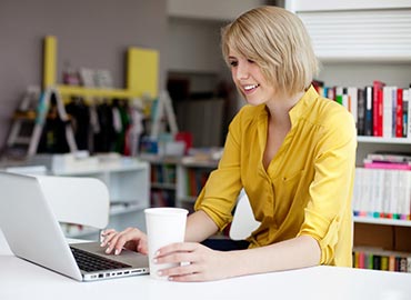 person21 Woman in yellow shirt working on laptop at desk with bookshelf behind her
