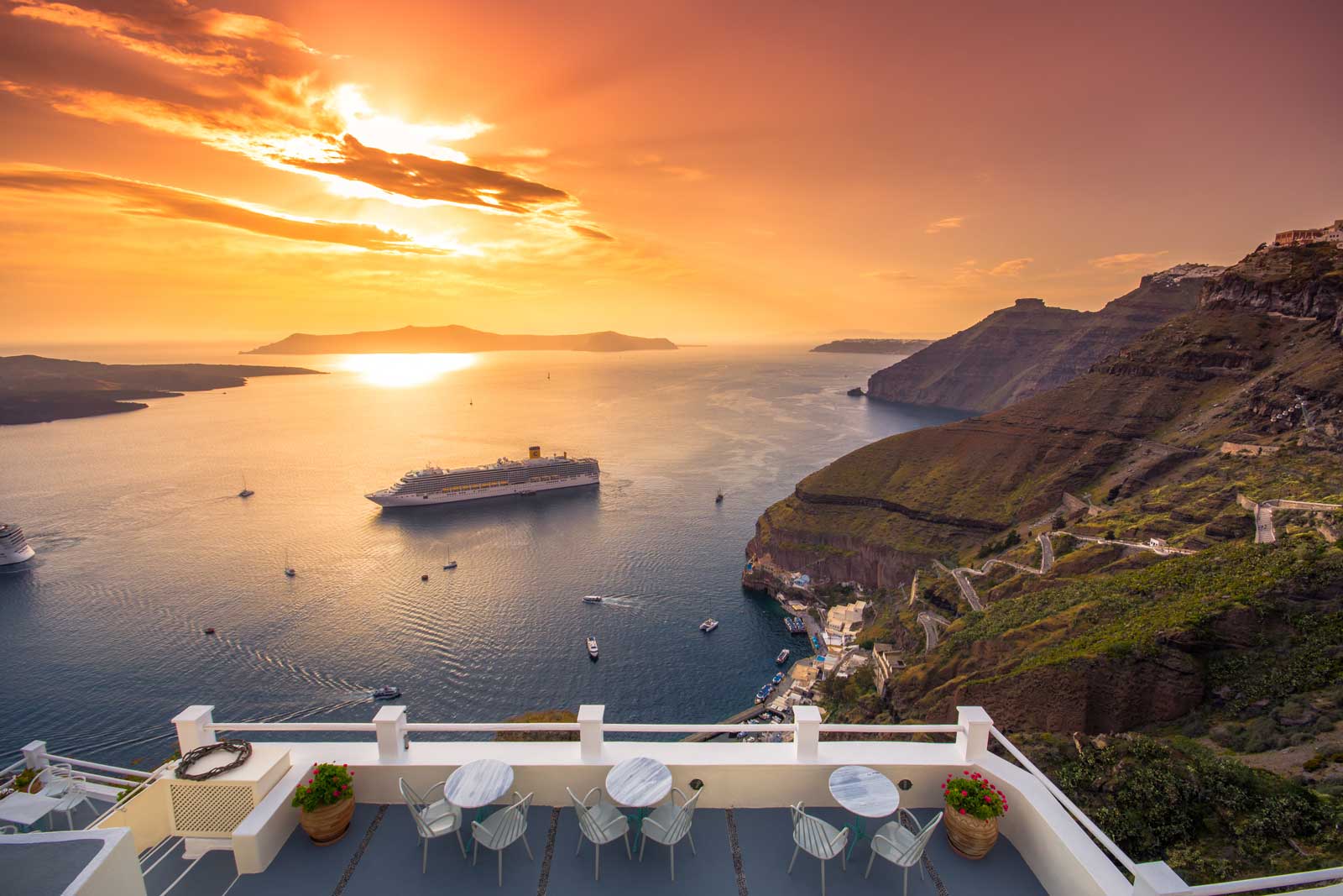 cruise-ship-santorini Cruise ship anchored in Santorini caldera at sunset, viewed from clifftop terrace with white railings
