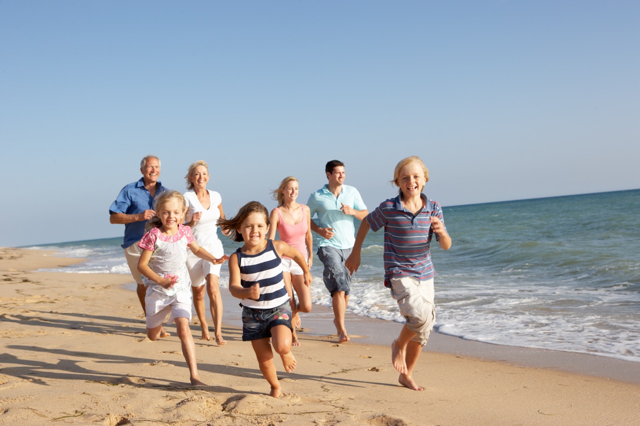 Portrait Of Three Generation Family On Beach Holiday Multi-generational family running together on sandy beach with ocean waves in background