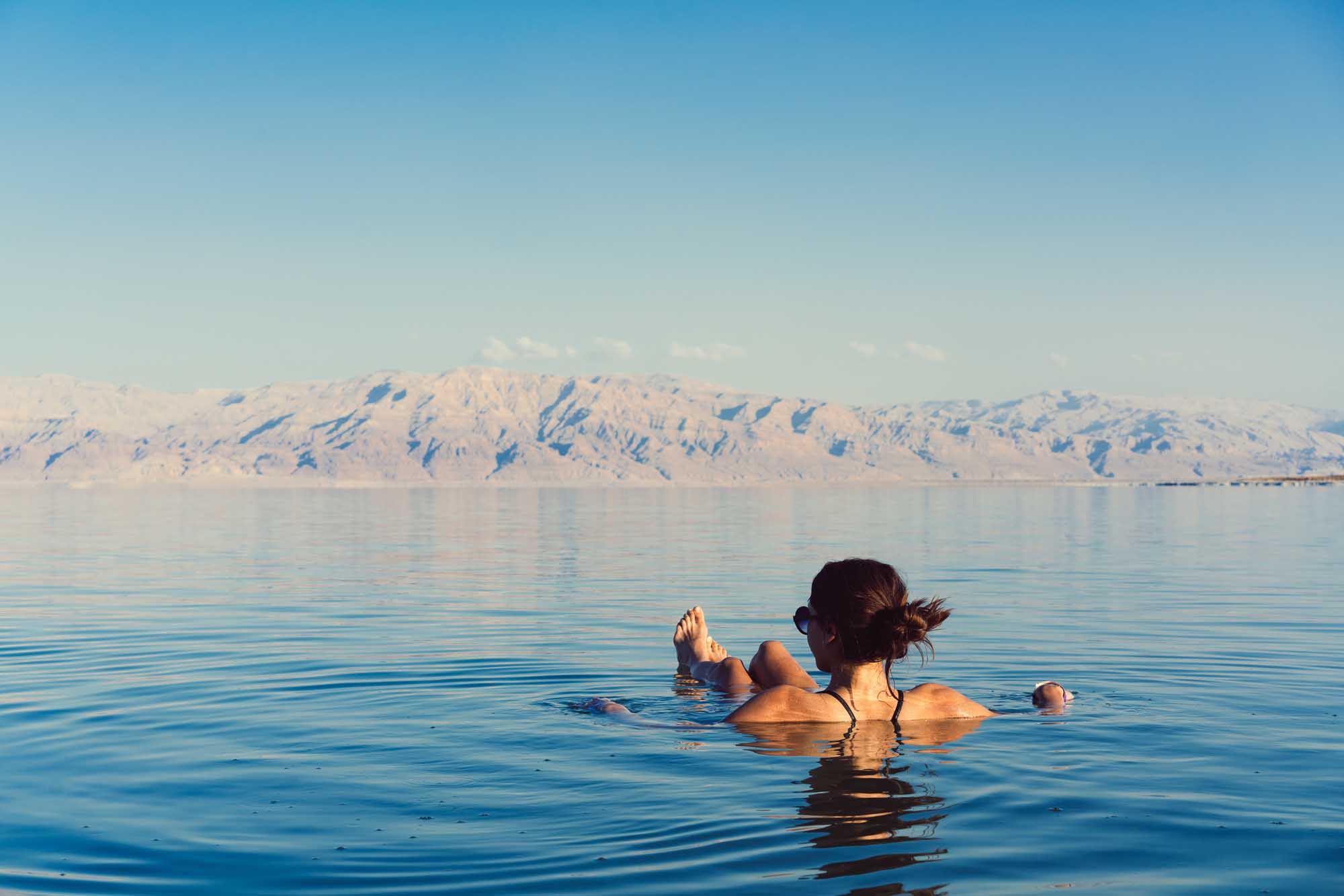 Swimming in the Dead Sea with Mountain Views A woman floats effortlessly in the crystal-clear waters of the Dead Sea, with snow-capped mountains